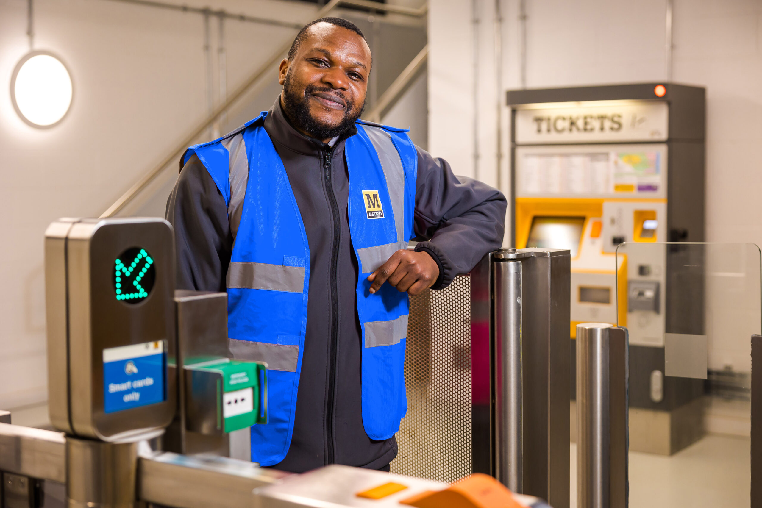 A man in a blue high vis vest stands at a Metro ticket gateline