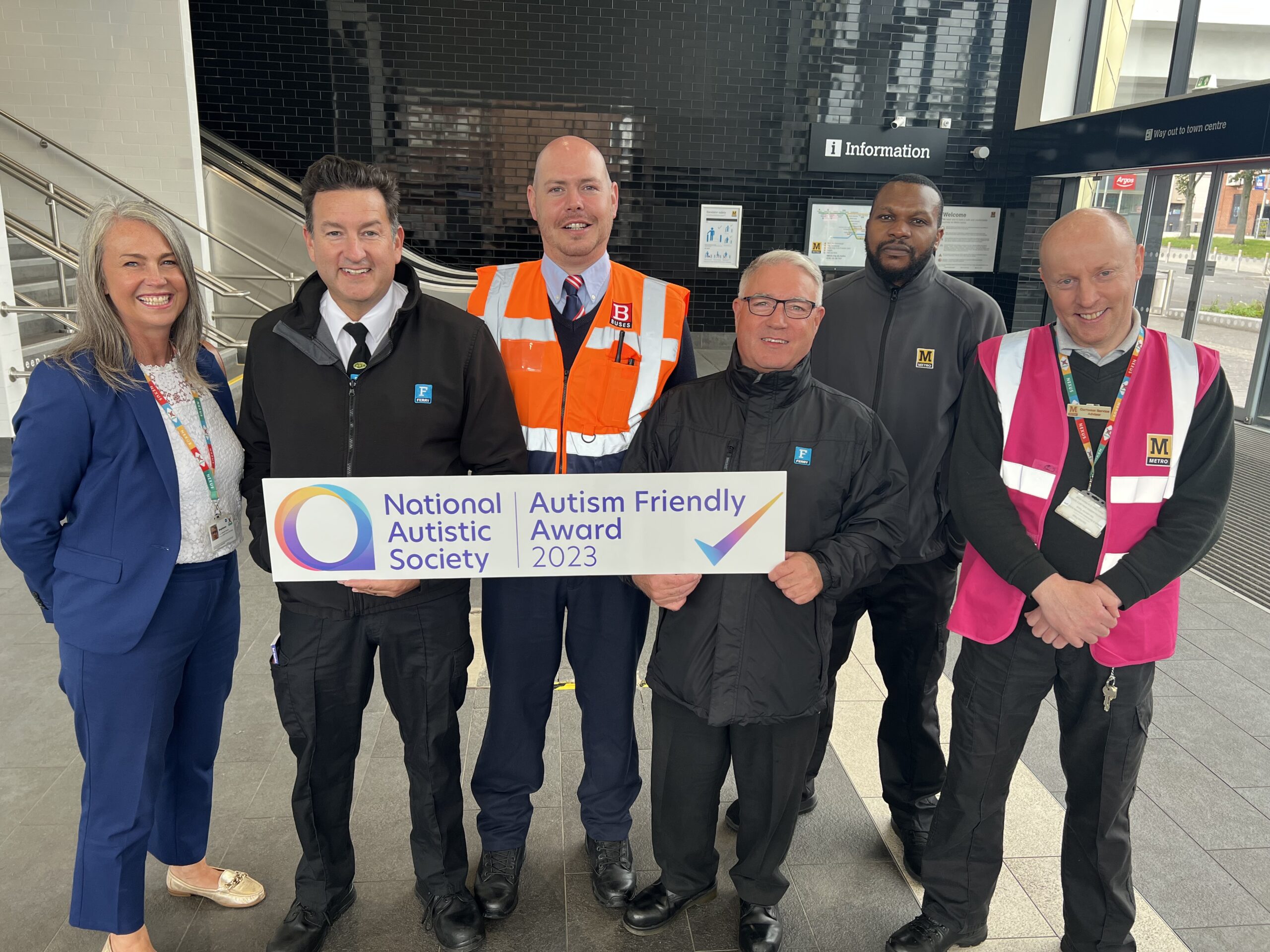 A group of Nexus employees in a Metro station with a sign saying Autism Friendly